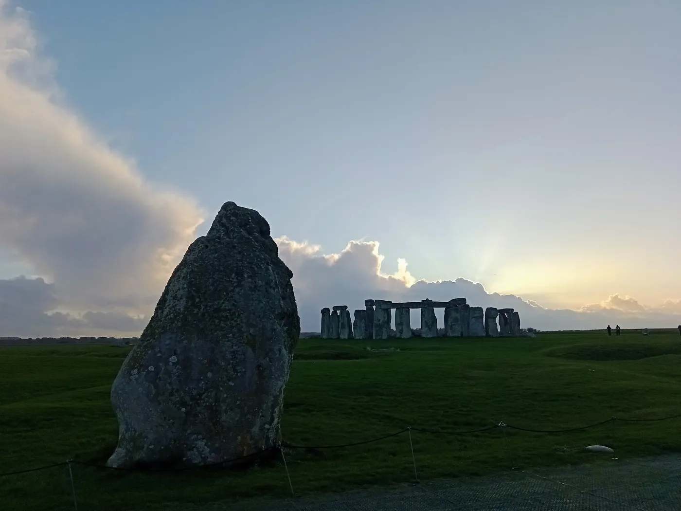 The Heel Stone in the foreground with the Stonehenge stone circle on the horizon under a cloudy evening sky